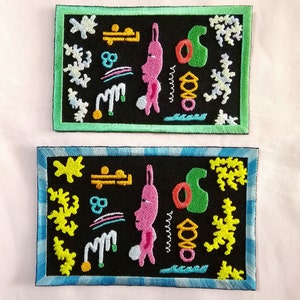 May include: Two embroidered patches with black backgrounds and colorful designs. The top patch has a green border and the bottom patch has a blue border. Both patches feature a pink creature with a long nose and a green object that resembles a leaf.