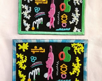 May include: Two embroidered patches with black backgrounds and colorful designs. The top patch has a green border and the bottom patch has a blue border. Both patches feature a pink creature with a long nose and a green object that resembles a leaf.