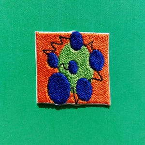 Può includere: Toppa ricamata con sfondo quadrato arancione e bordo bianco. La toppa presenta un centro verde con forme circolari blu e linee a zig-zag nere.