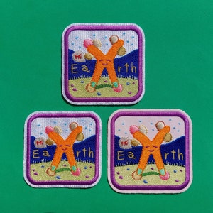 May include: Three embroidered patches with a square shape and a purple border. Each patch features an orange "X" character with a smiling face, pink accents, and the word "Earth". The background includes a blue mountain range and a light blue sky.