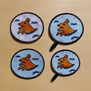 May include: Four embroidered iron-on patches featuring a cartoon brown dog swimming in blue water with white waves. The patches are circular and have a white background with blue trim. Two of the patches are attached to a black felt backing.