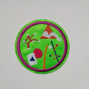 K&ouml;nnte beinhalten: Ein gr&uuml;ner und violetter gestickter Aufn&auml;her mit verschiedenen bunten Formen und Designs. Der Aufn&auml;her zeigt einen roten Kreis, ein blaues Dreieck, ein wei&szlig;es und orangefarbenes Zelt und einen wei&szlig;en und orangefarbenen Baum.