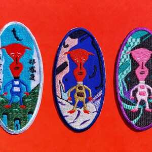 Op de afbeelding: Drie geborduurde patches met cartoonfiguren met verschillende gekleurde lichamen en achtergronden. De tekst op de linker patch luidt "Gaat het goed met je?" in het Japans.