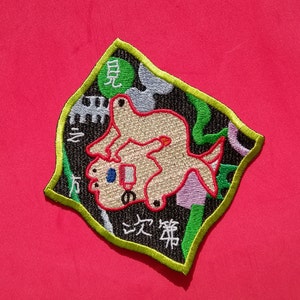 May include: Embroidered patch with a cartoon-style design featuring a light brown creature on top of another, with Japanese text. The patch has a black background with green, purple, and white accents, and a yellow-green border. The patch is on a red background.