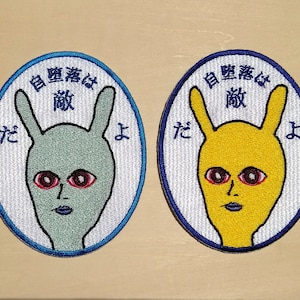 May include: Two embroidered patches with a white background and blue edging. The patches feature a cartoon bunny with red eyes and a blue mouth. The text on the patches reads "自意識は敵" which translates to "Self-consciousness is the enemy". One patch has a green bunny and the other has a yellow bunny.