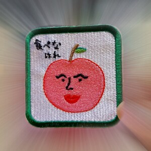 以下が含まれることがあります： 顔のある赤いリンゴが描かれた緑と白の刺繍パッチ。リンゴの上には緑の葉があり、黒い文字で「真好吃」と書かれている。