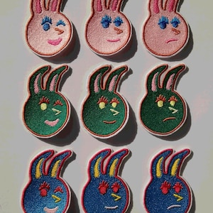 Könnte beinhalten: Neun gestickte Bügelbilder mit Hasengesichtern mit verschiedenen Ausdrücken. Die Hasen sind rosa, grün und blau mit gelben, weißen und roten Akzenten.