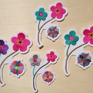 Peut inclure: Cinq patchs de fleurs brodés avec un fond en tissu blanc. Les fleurs sont de différentes couleurs, notamment rose, violet, turquoise et gris.