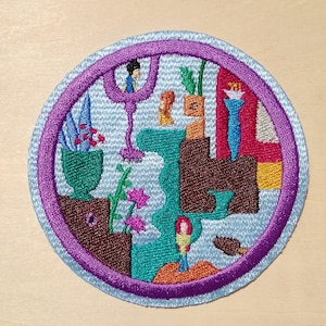 以下が含まれることがあります： 紫色の縁取りと青い背景を持つ円形の刺繍パッチ。パッチには、幾何学的な形、花、人物が描かれたカラフルな抽象的なデザインが施されています。