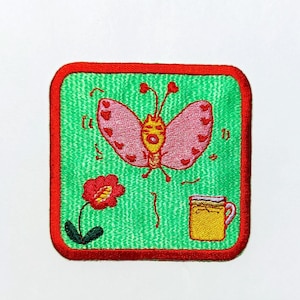 Puede incluir: Parche bordado con borde rojo, que presenta una mariposa rosa con marcas en forma de corazón, una flor roja y una taza amarilla sobre un fondo verde. El parche es de forma cuadrada.
