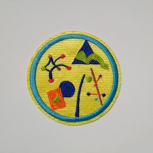 K&ouml;nnte beinhalten: Ein gelber gestickter Aufn&auml;her mit blauem Rand. Der Aufn&auml;her zeigt ein stilisiertes Design eines Berges, eines Baumes, eines Zeltes und anderer geometrischer Formen.