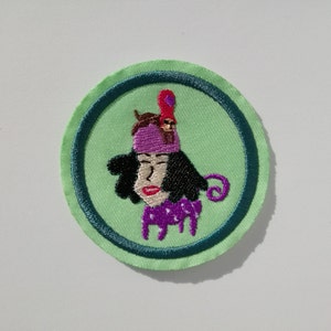 May include: A green and blue embroidered patch featuring a cartoon woman with a purple hat and a pink and red bird on top of her head.