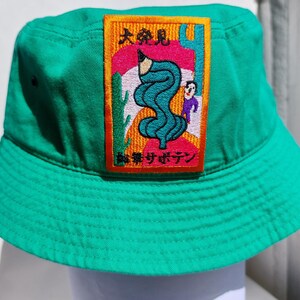 Pode incluir: Um chapéu bucket verde brilhante com um patch bordado colorido. O patch apresenta um lápis, um design azul ondulado, uma pessoa e texto japonês. O chapéu é feito de um tecido durável e tem um formato clássico.