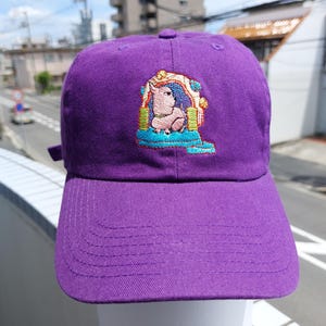 May include: A purple baseball cap with an embroidered design. The design features a seated cartoon pig in a colourful, ornate setting. The cap has a curved peak and an adjustable strap.