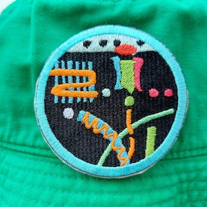 Pode incluir: Um chapéu de balde verde com um remendo redondo bordado colorido. O remendo apresenta um fundo preto com formas geométricas em laranja, azul, verde, rosa e branco.