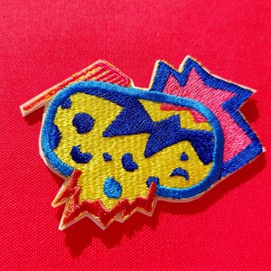Op de afbeelding: Geborduurde patch met een geel, blauw en rood ontwerp. De patch heeft een unieke vorm met een blauwe omtrek, een gele kern met blauwe stippen en rode en roze accenten. De patch staat op een rode achtergrond.