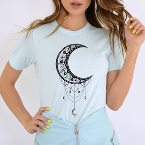 May include: A light blue t-shirt with a black crescent moon design. The moon is adorned with intricate details and has dangling chains with small circles.