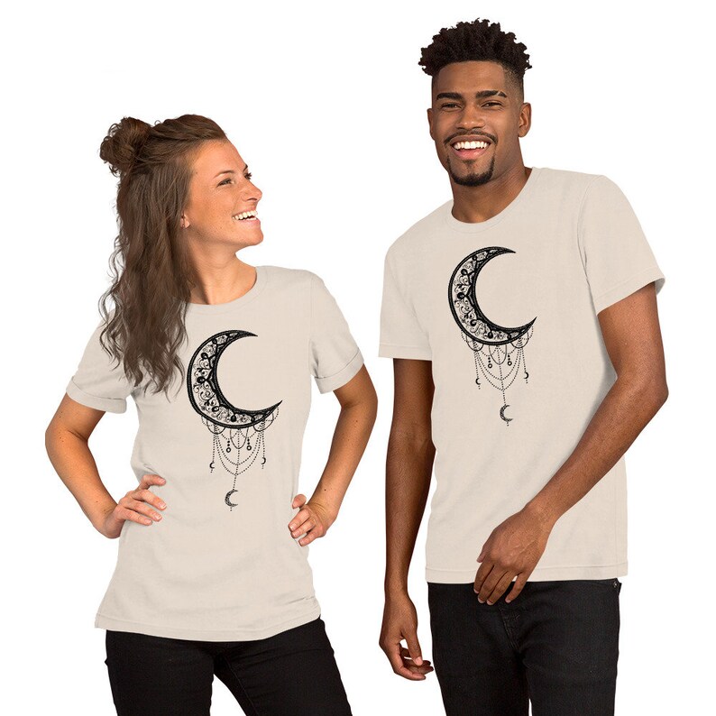 May include: Two people wearing beige t-shirts with a black crescent moon design. The moon is adorned with intricate details and dangling chains. The design is centered on the front of the shirts.