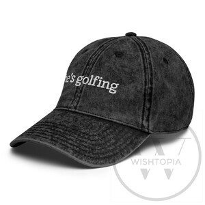 K&ouml;nnte beinhalten: Eine schwarze, ausgewaschene Baseballkappe mit dem wei&szlig;en Schriftzug "he's golfing". Die Kappe hat einen gebogenen Schirm und ein klassisches Sechs-Panel-Design. Der Hut ist im Vintage-Stil.