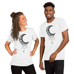 May include: Two white t-shirts with a black crescent moon design. The moon is adorned with intricate details and has dangling chains with small crescent moons at the end.