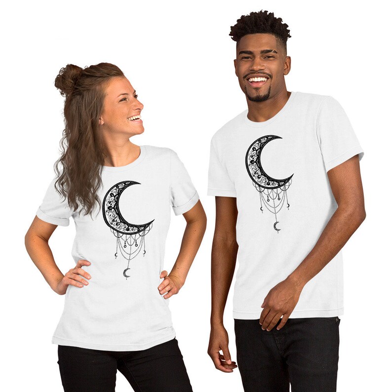 May include: Two white t-shirts with a black crescent moon design. The moon is adorned with intricate details and has dangling chains with small crescent moons at the end.