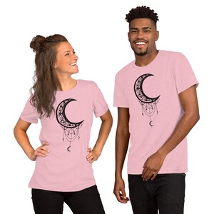 May include: Two pink t-shirts with a black crescent moon design. The moon is adorned with intricate details and has dangling chains with crescent moon charms.