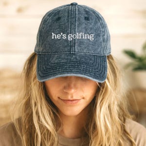 K&ouml;nnte beinhalten: Eine Baseballkappe in verwaschenem Denim-Blau mit dem wei&szlig;en, gestickten Text "he's golfing". Die Kappe hat einen gebogenen Schirm und einen Vintage-Look. Ein klassisches Accessoire f&uuml;r Outdoor-Aktivit&auml;ten.