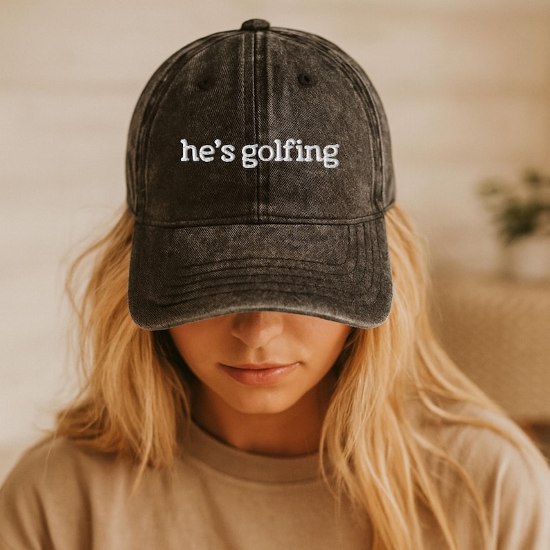 Vintage embroidered dad hat with “he’s golfing” text in white thread, washed black baseball cap styled on blonde woman, golf wife gift idea