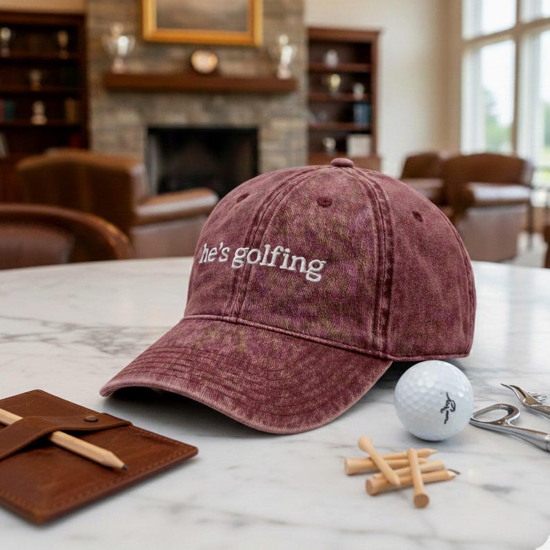 K&ouml;nnte beinhalten: Eine weinrote Baseballkappe mit dem wei&szlig;en Schriftzug "he's golfing". Ein Golfball, Holztees und ein kleines Ledernotizbuch mit einem Bleistift liegen ebenfalls auf dem Tisch. Der Hintergrund zeigt einen Kamin und einen Golfplatz.