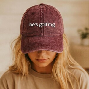 K&ouml;nnte beinhalten: Burgunderfarbene Baseballkappe mit dem gestickten Text "he's golfing" in Wei&szlig;. Die Kappe hat einen gebogenen Schirm und einen verblichenen Vintage-Look. Der Hut ist ein l&auml;ssiges Accessoire.
