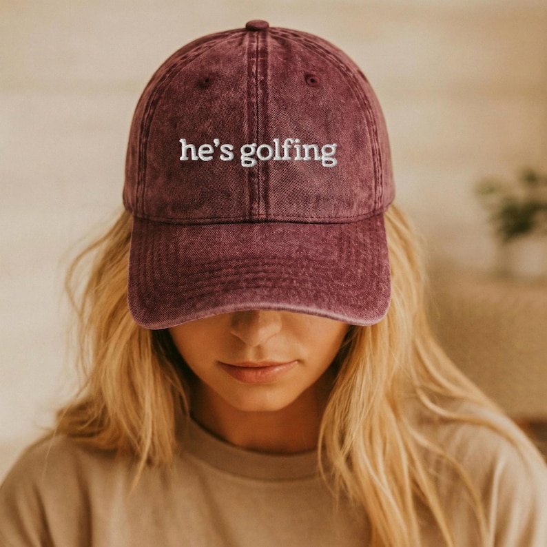 K&ouml;nnte beinhalten: Burgunderfarbene Baseballkappe mit dem gestickten Text "he's golfing" in Wei&szlig;. Die Kappe hat einen gebogenen Schirm und einen verblichenen Vintage-Look. Der Hut ist ein l&auml;ssiges Accessoire.