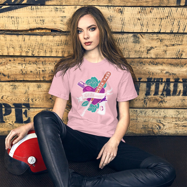 K&ouml;nnte beinhalten: Ein pinkfarbenes T-Shirt mit einem Aufdruck eines Herzens mit einem Lippenstiftkuss, einem Baseballschl&auml;ger, einem Rasiermesser und einer Rose. Der Text "Bad Influence" ist auf einem Banner geschrieben.