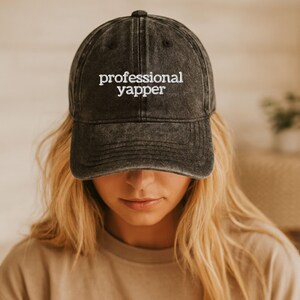 May include: A dark gray baseball cap with the embroidered text "professional yapper" in white. The cap is worn by a person with blonde hair and a neutral-colored shirt.