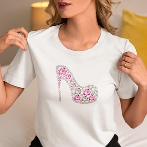 Könnte beinhalten: Weißes T-Shirt mit einem Design einer High-Heel-Schuh. Der Schuh ist mit rosa und klaren Strasssteinen verziert, was einen funkelnden Effekt erzeugt. Der Absatz besteht aus einer Reihe von Strasssteinen. Ein lässiges Kleidungsstück für den Alltag.
