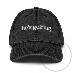 K&ouml;nnte beinhalten: Schwarze Baseballkappe im Vintage-Stil mit dem gestickten Text "he's golfing" in Wei&szlig;. Die Kappe hat einen gebogenen Schirm und einen Knopf oben. Der Stoff hat einen verblichenen, abgenutzten Look.