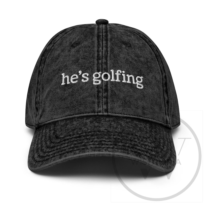 K&ouml;nnte beinhalten: Schwarze Baseballkappe im Vintage-Stil mit dem gestickten Text "he's golfing" in Wei&szlig;. Die Kappe hat einen gebogenen Schirm und einen Knopf oben. Der Stoff hat einen verblichenen, abgenutzten Look.