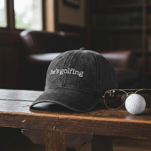 K&ouml;nnte beinhalten: Eine schwarze Baseballkappe mit dem wei&szlig;en Schriftzug "he's golfing". Die Kappe liegt auf einem Holztisch neben einem Golfball und einer goldgerahmten Sonnenbrille. Der Hintergrund ist unscharf, was auf eine Innenaufnahme hindeutet.