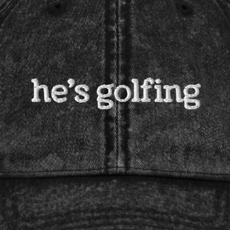 K&ouml;nnte beinhalten: Eine schwarze Baseballkappe aus Denim mit dem wei&szlig;en gestickten Text "he's golfing". Die Kappe hat einen gebogenen Schirm und ein verwaschenes Aussehen, was einen Vintage- oder l&auml;ssigen Stil suggeriert.