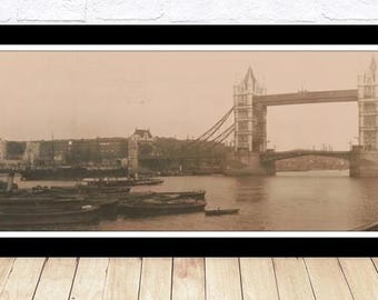 Può includere: Una fotografia panoramica in bianco e nero del Tower Bridge a Londra, in Inghilterra. Il ponte è un ponte sospeso con due torri e una passerella. Il ponte è sopra un fiume con barche e navi nell'acqua. La fotografia è incorniciata in una cornice nera.
