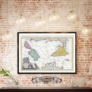 Op de afbeelding: Een vintage kaart van de Middellandse Zee met eilanden die in roze, groen en geel zijn gemarkeerd. De kaart is zwart ingelijst en hangt aan een bakstenen muur met meerdere bolvormige lampen.