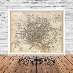 Op de afbeelding: Een ingelijste vintage kaart van Birmingham, Engeland, met een gedetailleerde plattegrond van straten, gebouwen en groene ruimtes. De kaart zit in een witte lijst, tegen een witte bakstenen muur en een houten vloer. De tekst "XXLPRINTS" staat onderaan.