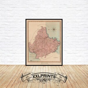 Puede incluir: Mapa vintage enmarcado de Devonshire, Inglaterra, en un marco negro. El mapa presenta un diseño detallado en rosa y beige con una rosa de los vientos y texto. El mapa se muestra contra una pared de ladrillo blanco y un suelo de madera.