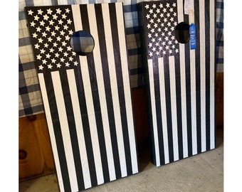 May include: Two black and white American flag cornhole boards. The boards are made of wood and have a black and white striped design. The boards are leaning against a wall.