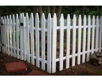 May include: A white picket fence with pointed tops surrounds a grassy area. The fence is made of individual wooden slats that are joined together to create a continuous barrier. The fence is in good condition and appears to be well-maintained.