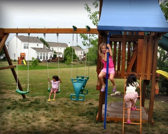 May include: Three children play on a wooden playground set with a blue canopy. Two girls are on a swing set and one girl is climbing a ladder. The playground set is in a backyard with green grass and houses in the background.