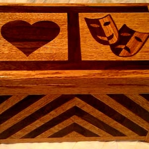 Puede incluir: Una caja de madera con un patr&oacute;n de chevron en la parte inferior y un coraz&oacute;n y dos m&aacute;scaras de teatro en la parte superior. La caja est&aacute; hecha de madera clara y oscura.