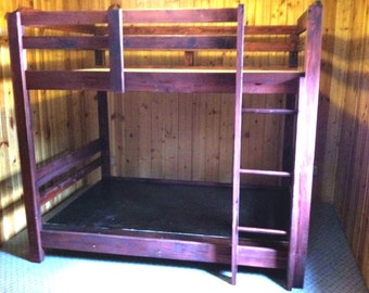 May include: A wooden bunk bed with a dark reddish-brown stain. The bed has a ladder for access to the top bunk and safety rails. The frame is made of thick wooden beams, and the bed has a simple, rustic design.