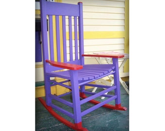 May include: Purple rocking chair with red accents. The chair is made of wood and has a slatted back and seat. The chair is sitting on a wooden porch.