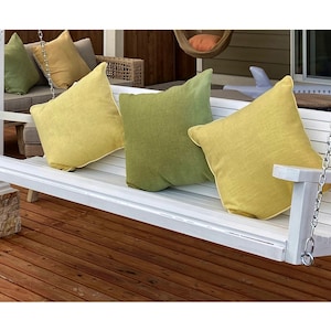 May include: White wooden porch swing with a white metal chain hanging from a wooden pergola. The swing is decorated with four throw pillows, two yellow and two green.