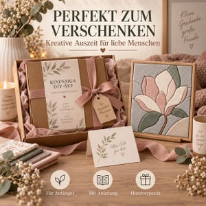 Könnte beinhalten: Ein Kinusaiga DIY-Set in einem braunen Karton mit altrosa Schleife, bestehend aus einem fertigen Stoffbild mit rosa-grünem Magnolienmotiv, einer Grußkarte mit Blattillustrationen sowie mehreren gefalteten Stoffstücken in verschiedenen Farben.